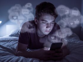 Adolescente sozinho em quarto escuro olhando para a tela do celular, cercado por ícones de redes sociais, simbolizando o impacto das redes na saúde mental dos jovens.