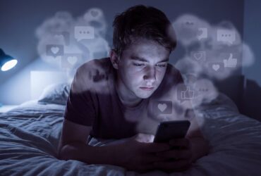 Adolescente sozinho em quarto escuro olhando para a tela do celular, cercado por ícones de redes sociais, simbolizando o impacto das redes na saúde mental dos jovens.