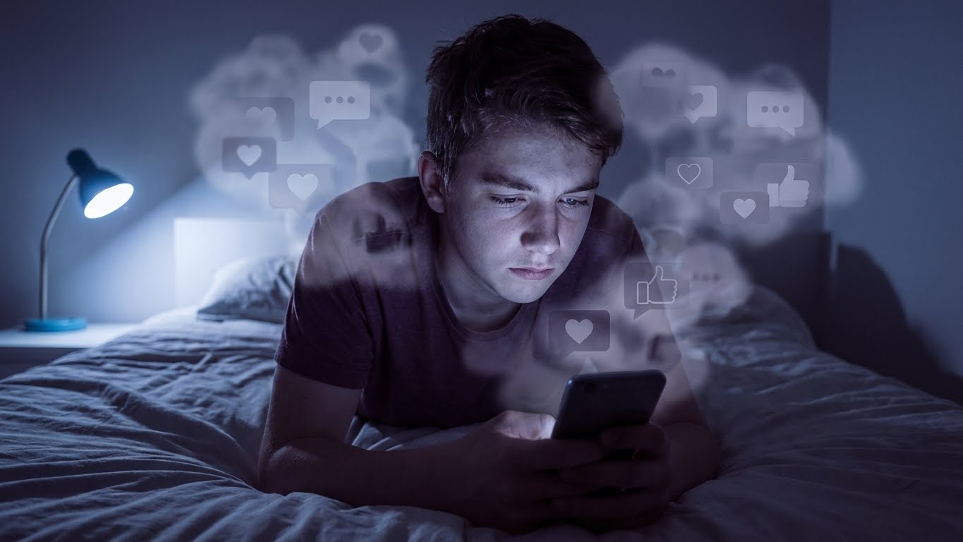 Adolescente sozinho em quarto escuro olhando para a tela do celular, cercado por ícones de redes sociais, simbolizando o impacto das redes na saúde mental dos jovens.